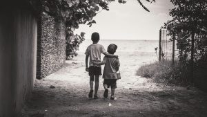 grayscale beach scene with two boys walking in the sand towards the water with leaves overhead and a building to the side