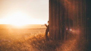 man playing a trumpet, leaning against a wooden building and looking out at a golden field