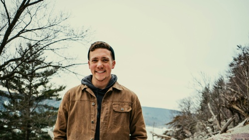 a smiling young man standing outdoors with a mountainous background