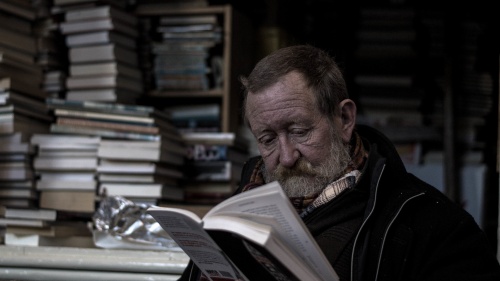 An older man with a beard reading a book. Stacks of books are behind him.