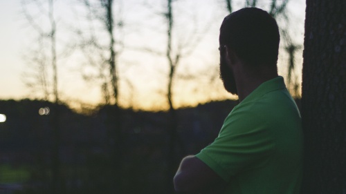 A man with arms crossed leaning against a tree looking at the sun setting.