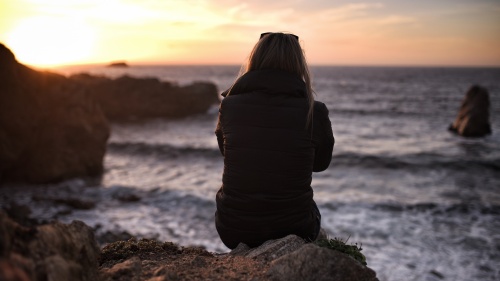 A young woman sitting on an rock looking at the sun setting over the ocean.