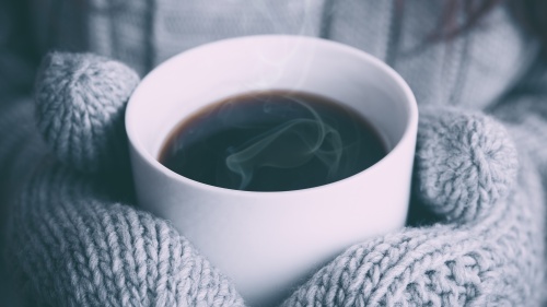 A person wearing mittens holding a hot beverage with steam rising.