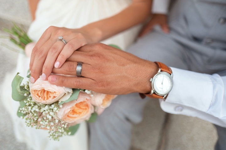 a married couple's hands layered over a pink bouquet of flowers and wearing wedding rings