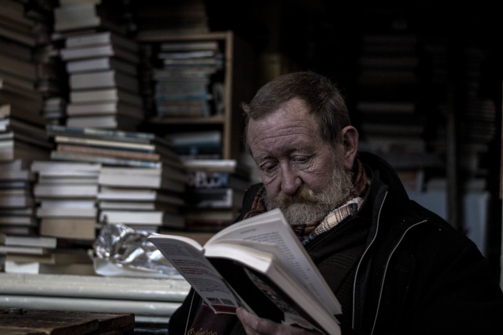 An older man with a beard reading a book. Stacks of books are behind him.