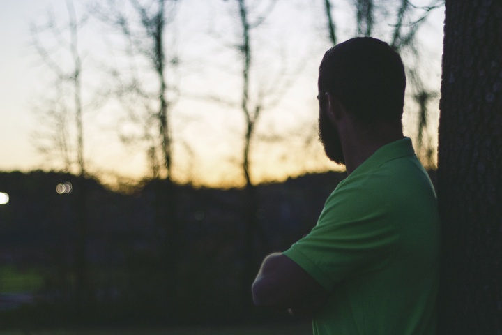 A man with arms crossed leaning against a tree looking at the sun setting.