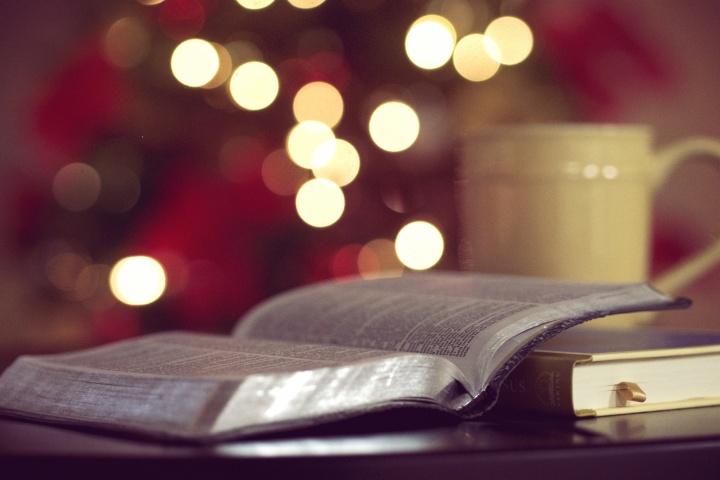 An opened Bible in the foreground and a Christmas tree lit up in the background.