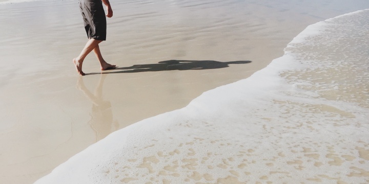 A man walking on the beach beside the ocean wave. His shadow on the flat sand.