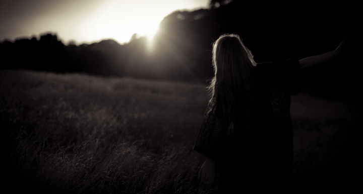 A woman standing in a field with the sun glaring behind a row of trees.