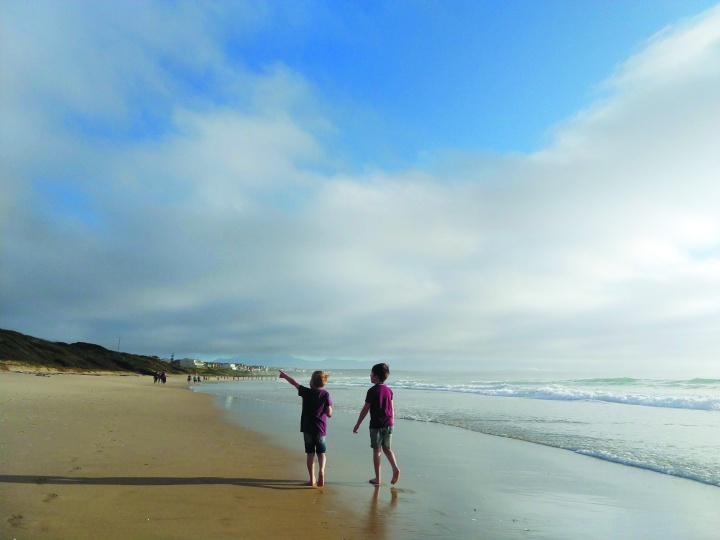 Two young boys walking along a beach, one pointing at something in the distance to the other. Wispy clouds cover part of the bright blue sky!