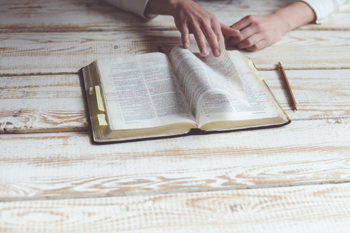 Photo of a marked up bible on a wooden table with a person's hands flipping through it. A pencil lays on the table next to the Bible.